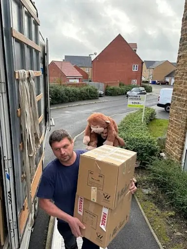 Gentlevan mover carrying a fragile-marked carton and a child's soft toy out to the van, sold board on the drive behind