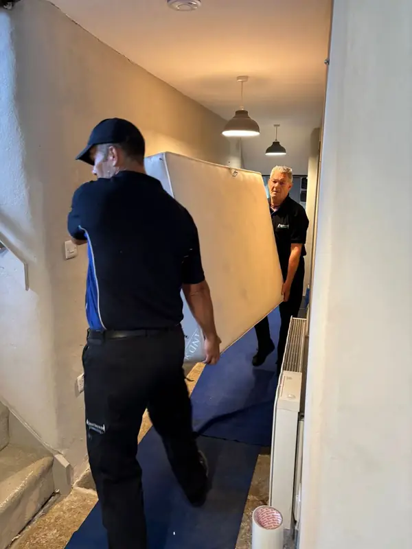 Two removal crew in Gentlevan polo shirts carrying a mattress along a narrow hallway over a blue carpet protection runner