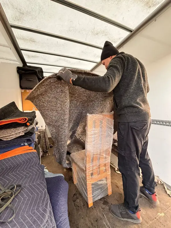 Removal crew member unfolding a padded moving blanket inside a removal van beside a bubble-wrapped chair and folded blankets