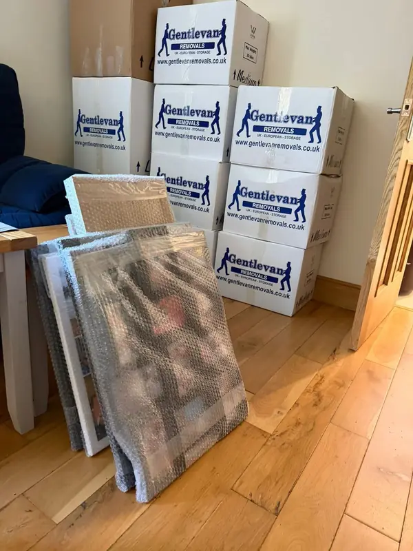 Stacked Gentlevan branded removal cartons beside bubble-wrapped framed pictures on a wooden floor