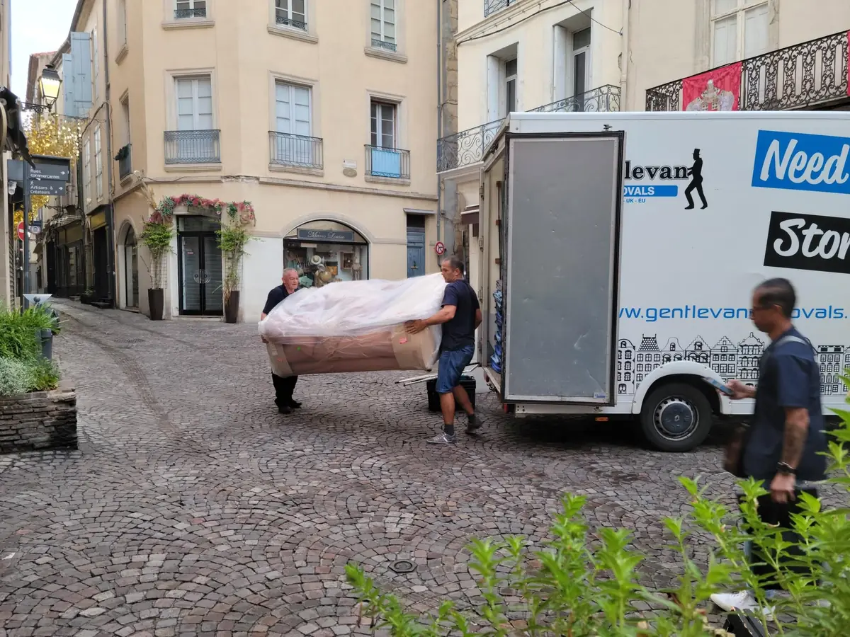Two Gentlevan Removals crew carrying a wrapped item across a cobbled street to a smaller box vehicle in a French town centre