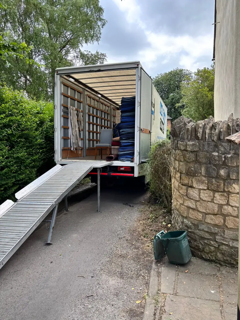 A Gentlevan Removals lorry with the tail lift and loading ramp down in a narrow lane beside a stone wall, blankets stacked inside
