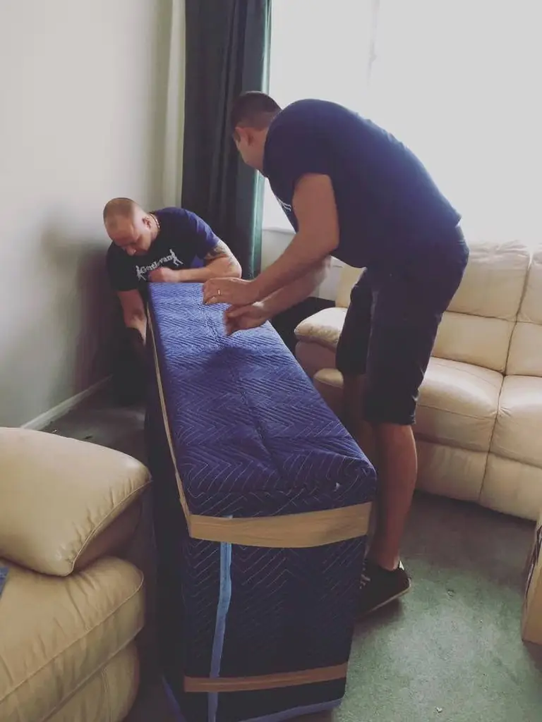 Two Gentlevan Removals crew fitting a padded quilted cover over a mattress in a lounge before loading