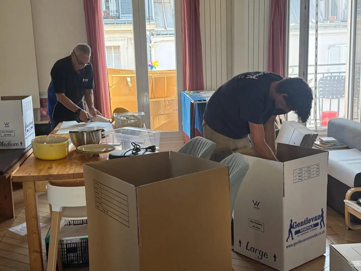 Two Gentlevan Removals crew packing kitchenware at a dining table in an apartment with tall windows, with branded cartons ready to fill