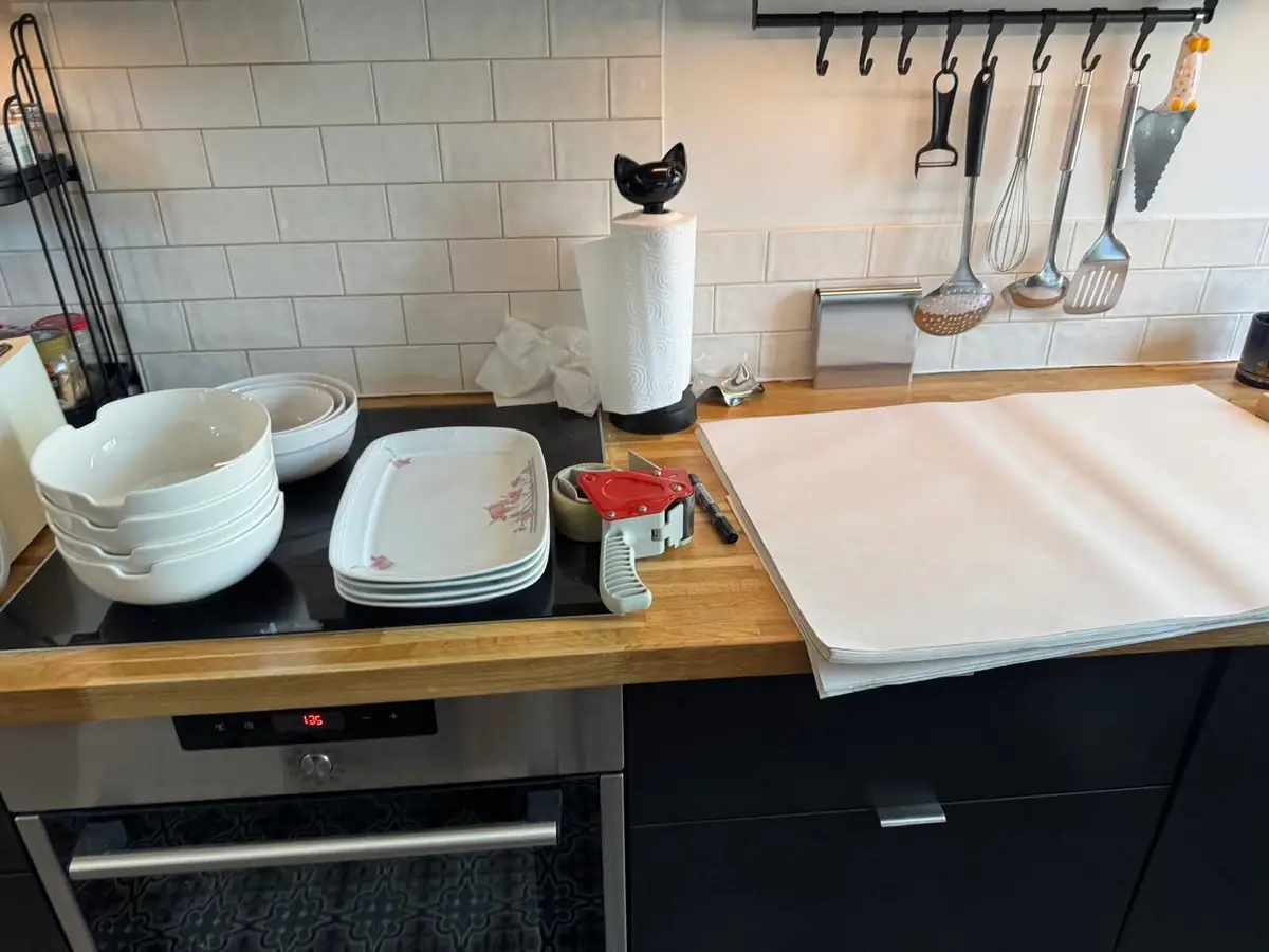 Stacked white bowls and serving platters on a hob beside a tape gun, a marker pen and a pile of white packing paper on a wooden worktop