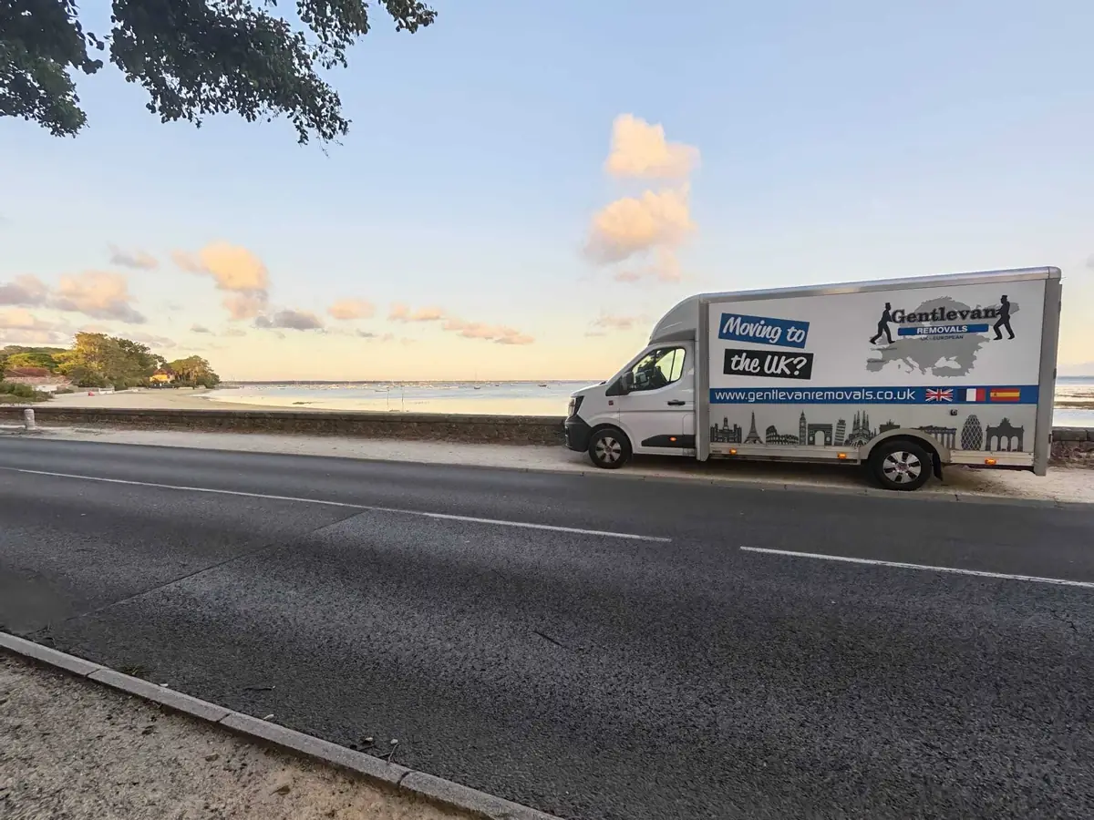 Gentlevan Removals van signwritten Moving to the UK parked on a coast road beside a bay with moored boats at sunset